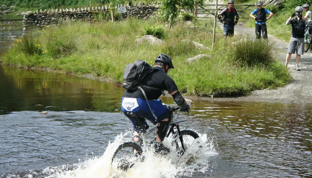 Mountain biker splashing through a shallow river while fellow cyclists watch from a rural track