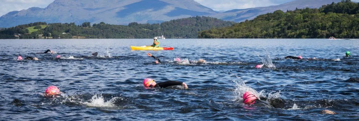 Open-water swimmers in pink caps racing across a mountain lake, accompanied by a kayaker.