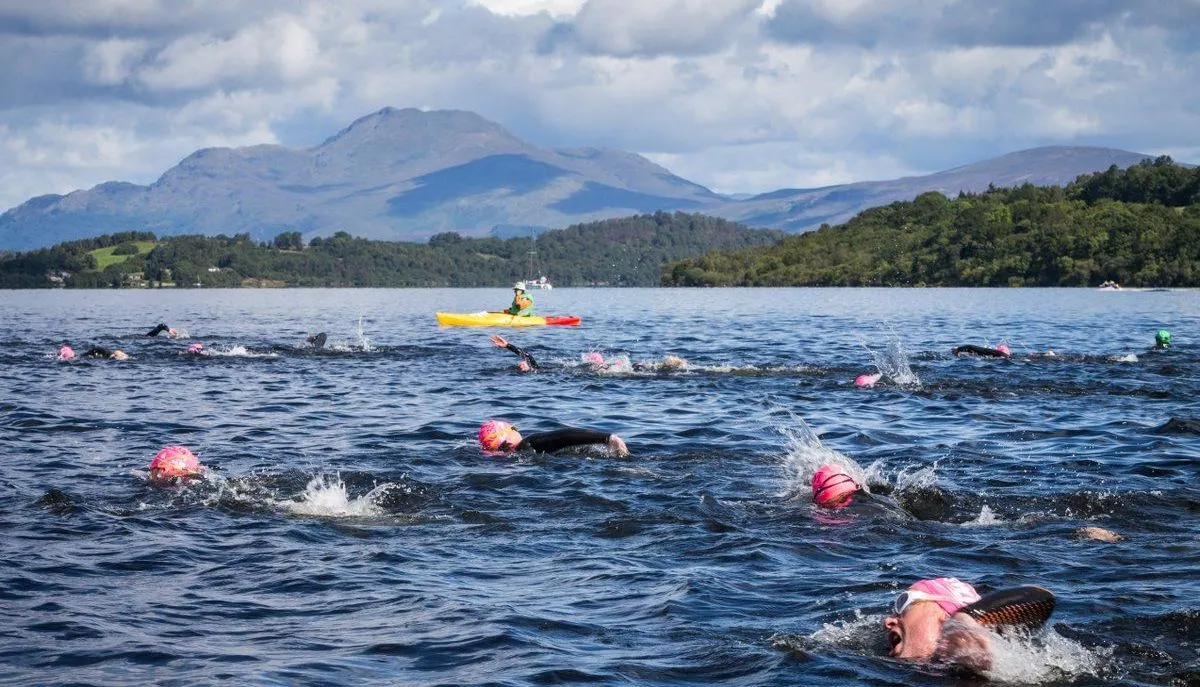 Open-water swimmers in pink caps racing across a mountain lake, accompanied by a kayaker.