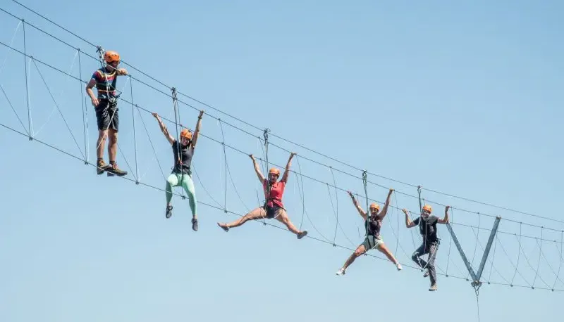 Five helmeted climbers cross a high rope bridge at Honister Slate Mine against a clear blue sky.