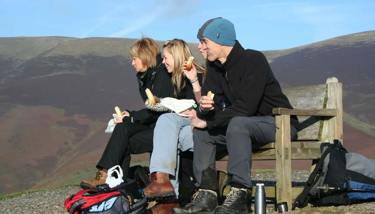 Three hikers eating sandwiches on a wooden bench overlooking rolling hills