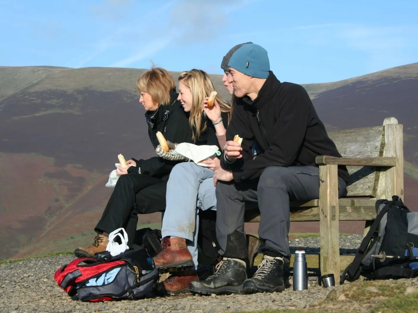 Three hikers eating sandwiches on a wooden bench overlooking rolling hills