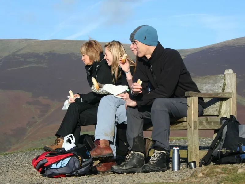 Three hikers eating sandwiches on a wooden bench overlooking rolling hills