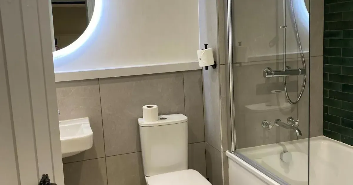 Modern compact bathroom with toilet, bathtub shower, glass screen and illuminated circular mirror.