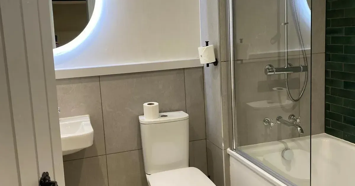 Modern compact bathroom with toilet, bathtub shower, glass screen and illuminated circular mirror.