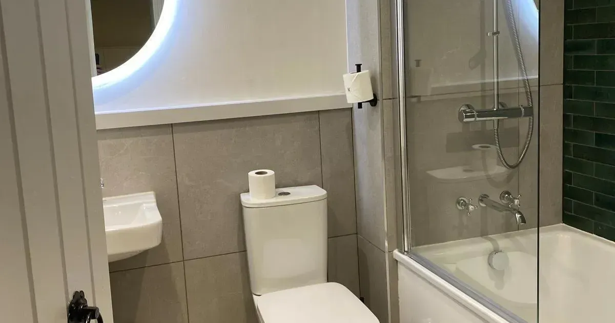 Modern compact bathroom with toilet, bathtub shower, glass screen and illuminated circular mirror.