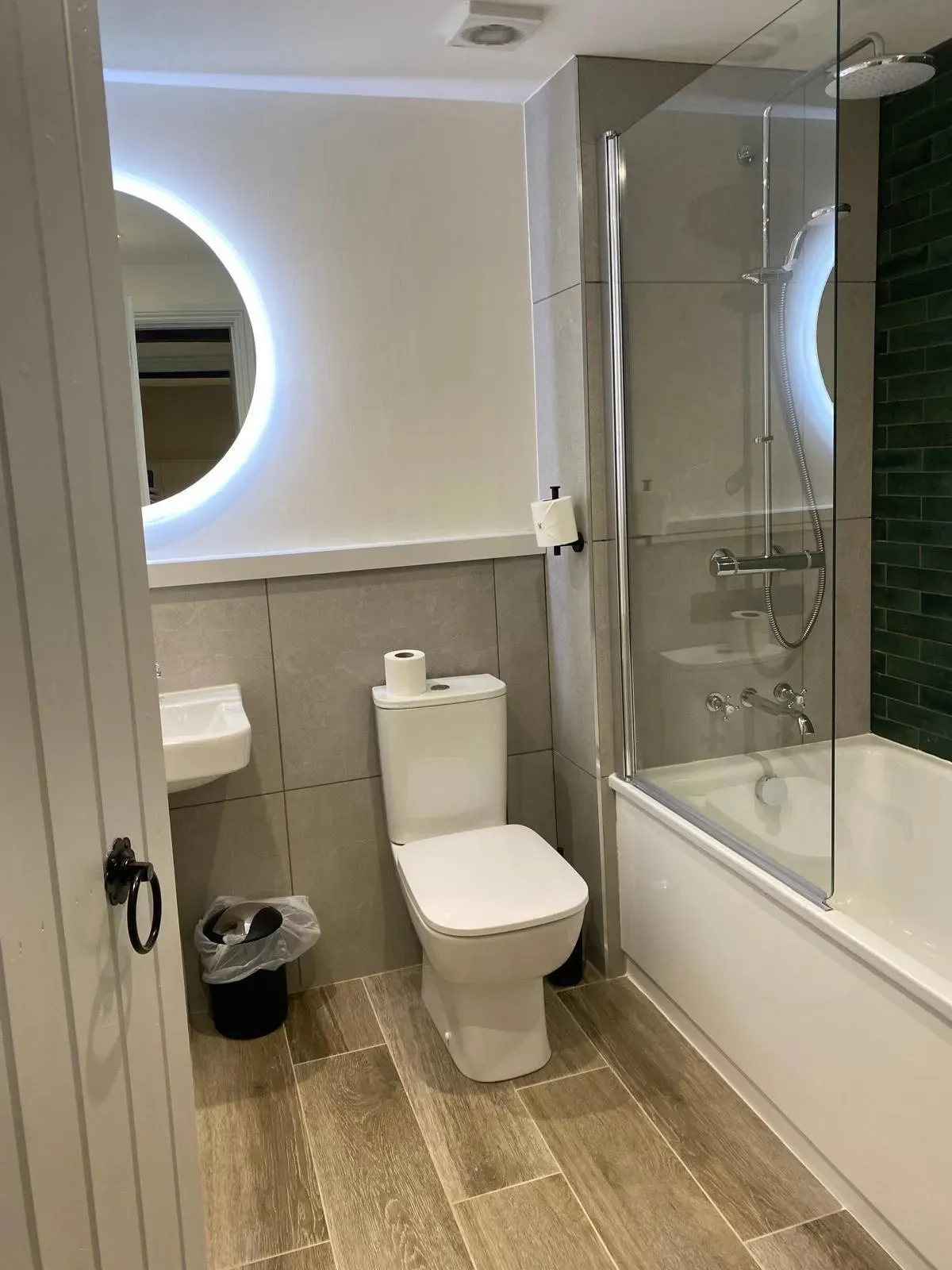 Modern compact bathroom with toilet, bathtub shower, glass screen and illuminated circular mirror.