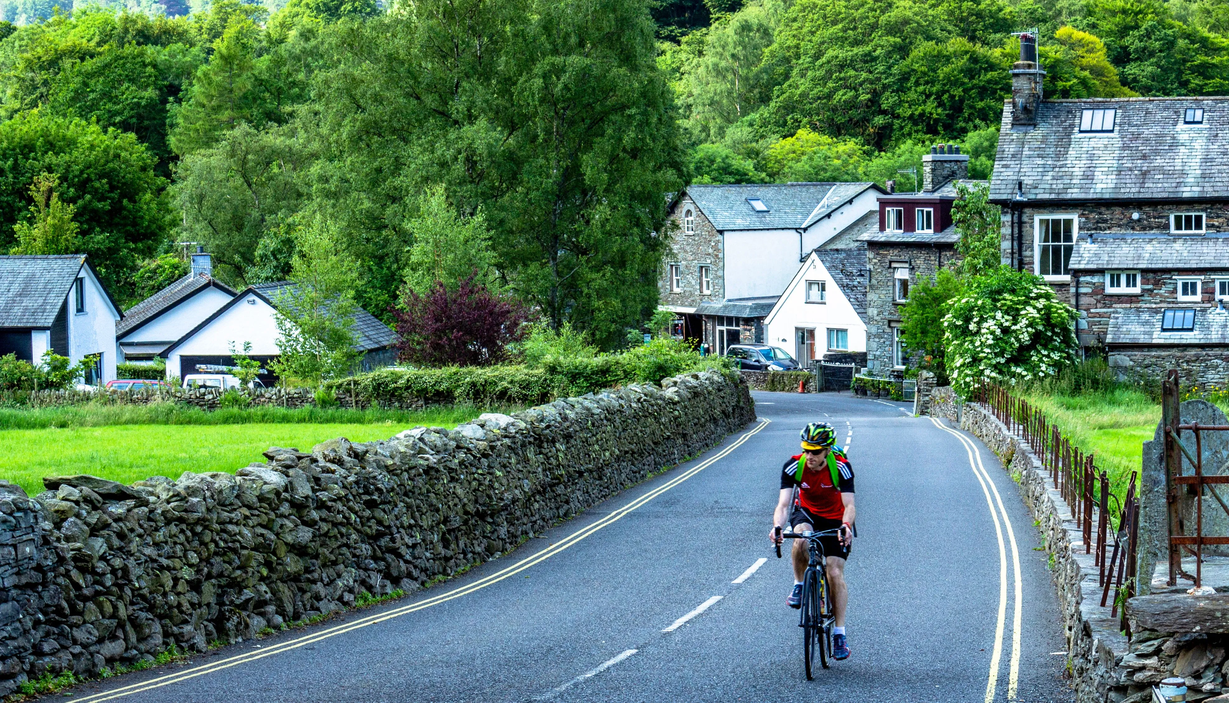 Cyclist riding along a winding country road through a green village with stone cottages and dry-stone walls.