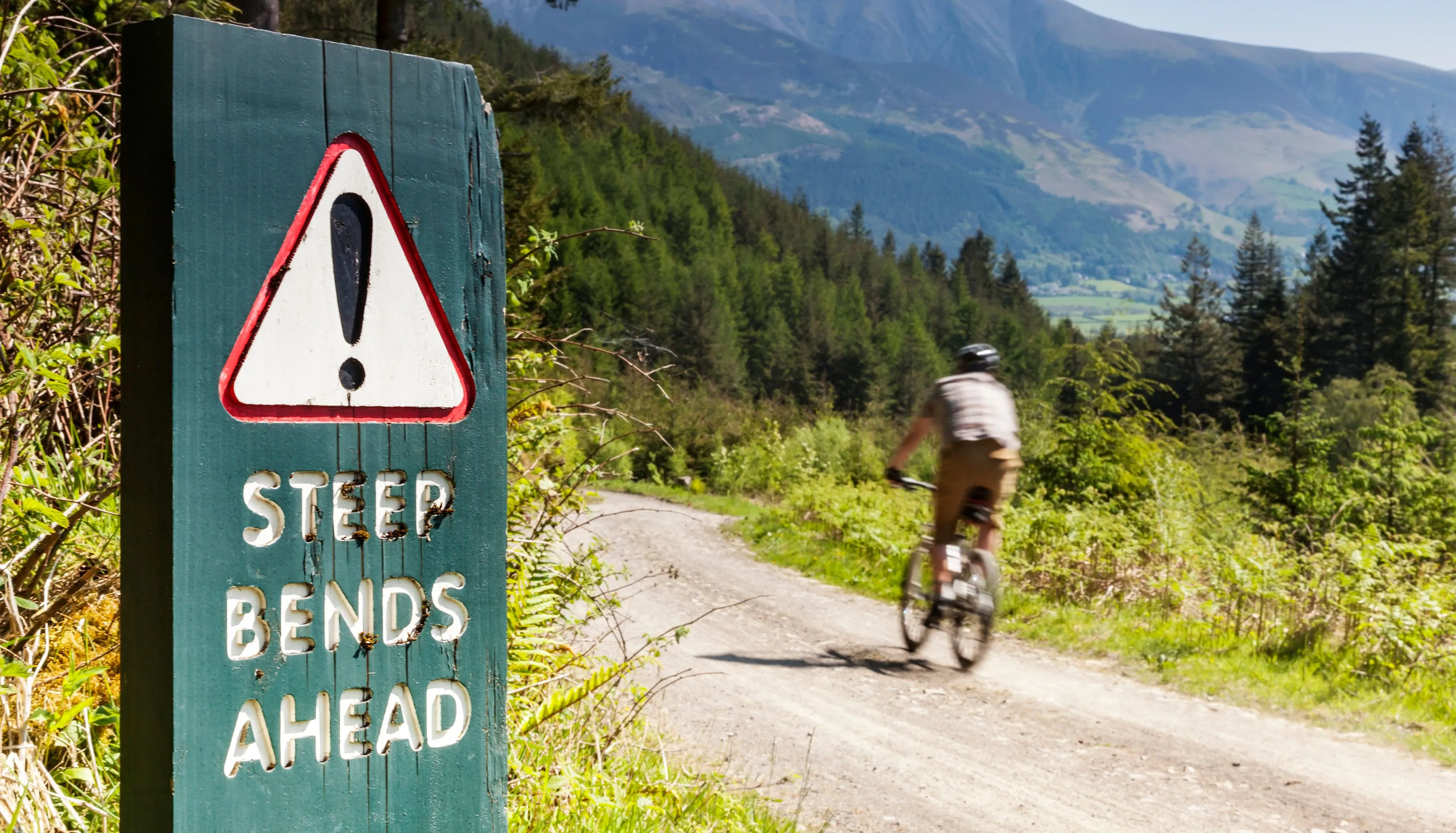Warning sign reading “Step bends ahead” beside a mountain bike trail in a forested valley.
