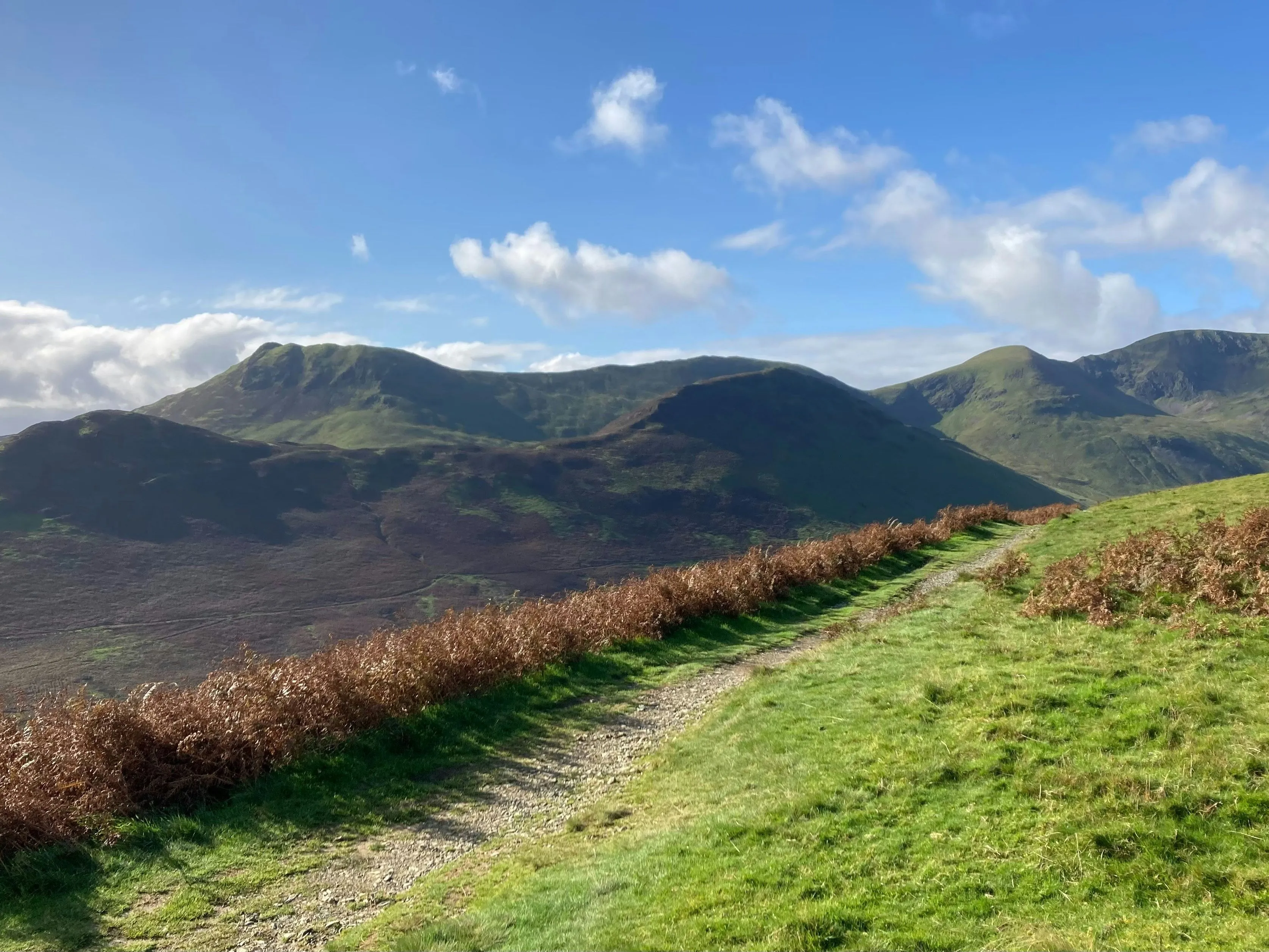 Grassy mountain path overlooking green hills beneath a bright blue sky with scattered clouds