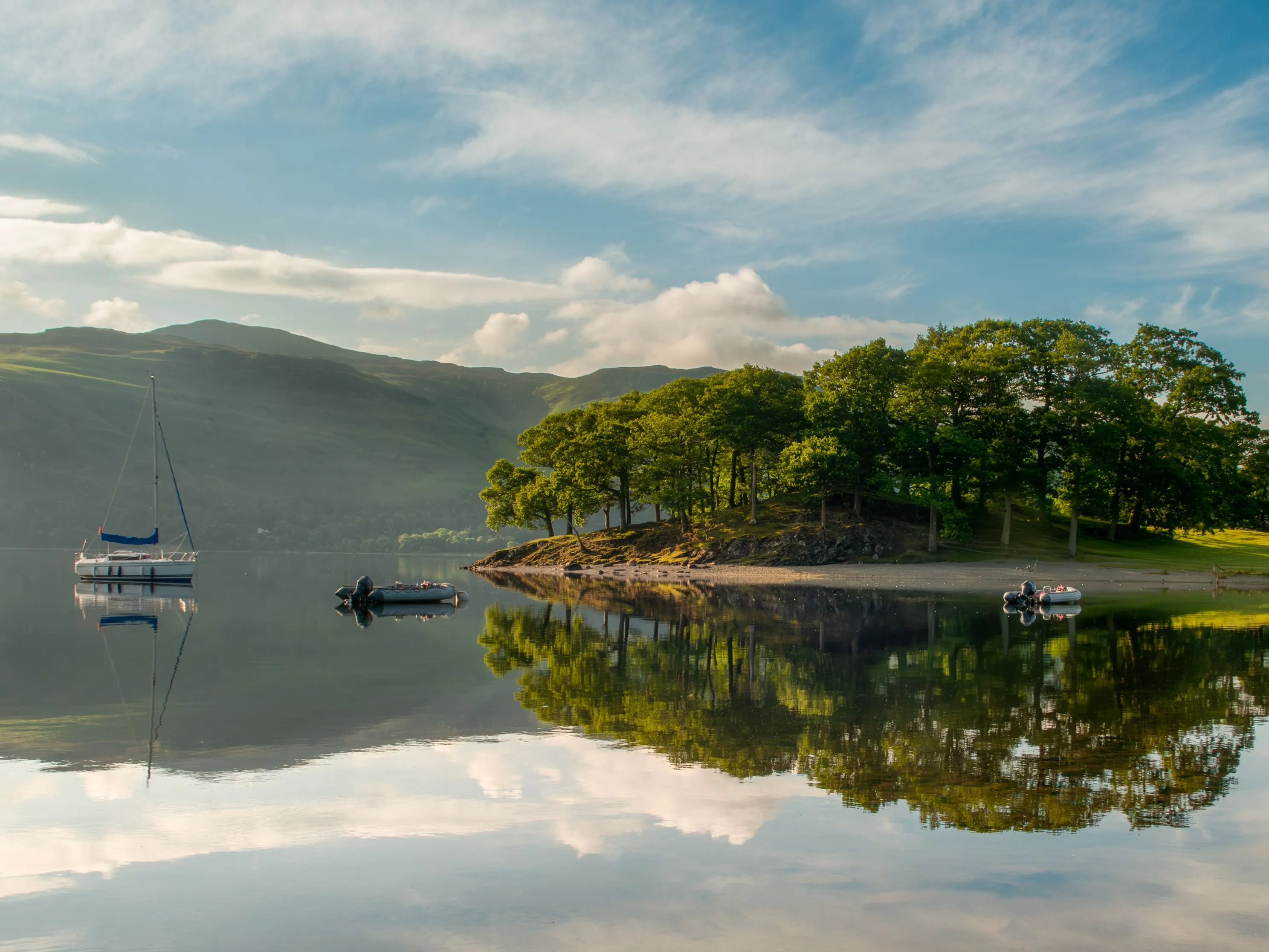 Sailing boat and small dinghies on a calm lake beside a tree-covered shore and hills, reflected in the water.