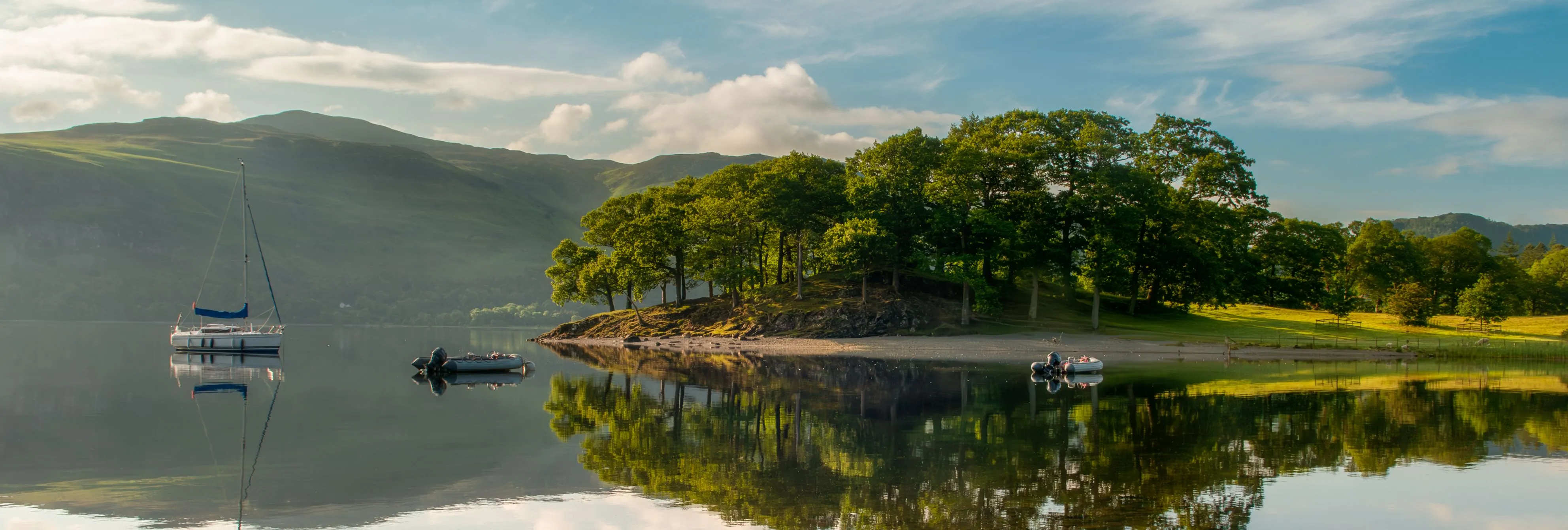Sailing boat and small dinghies on a calm lake beside a tree-covered shore and hills, reflected in the water.