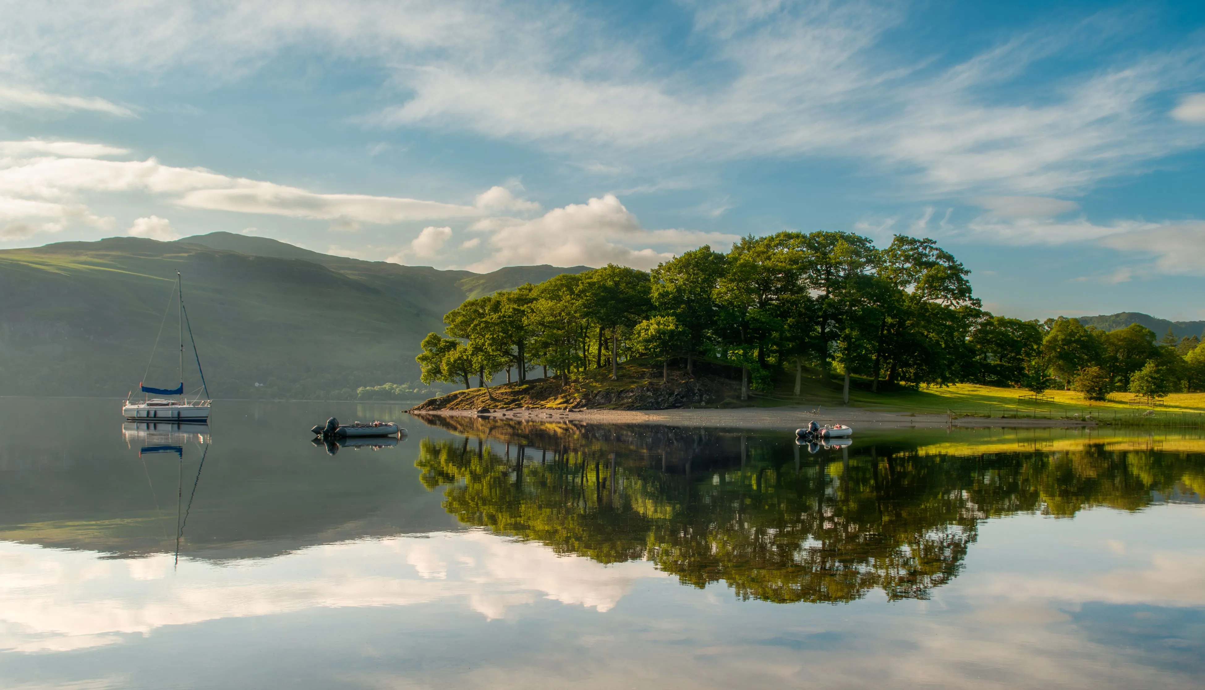 Sailing boat and small dinghies on a calm lake beside a tree-covered shore and hills, reflected in the water.