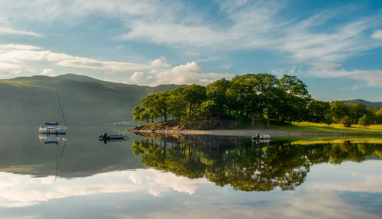Sailing boat and small dinghies on a calm lake beside a tree-covered shore and hills, reflected in the water.