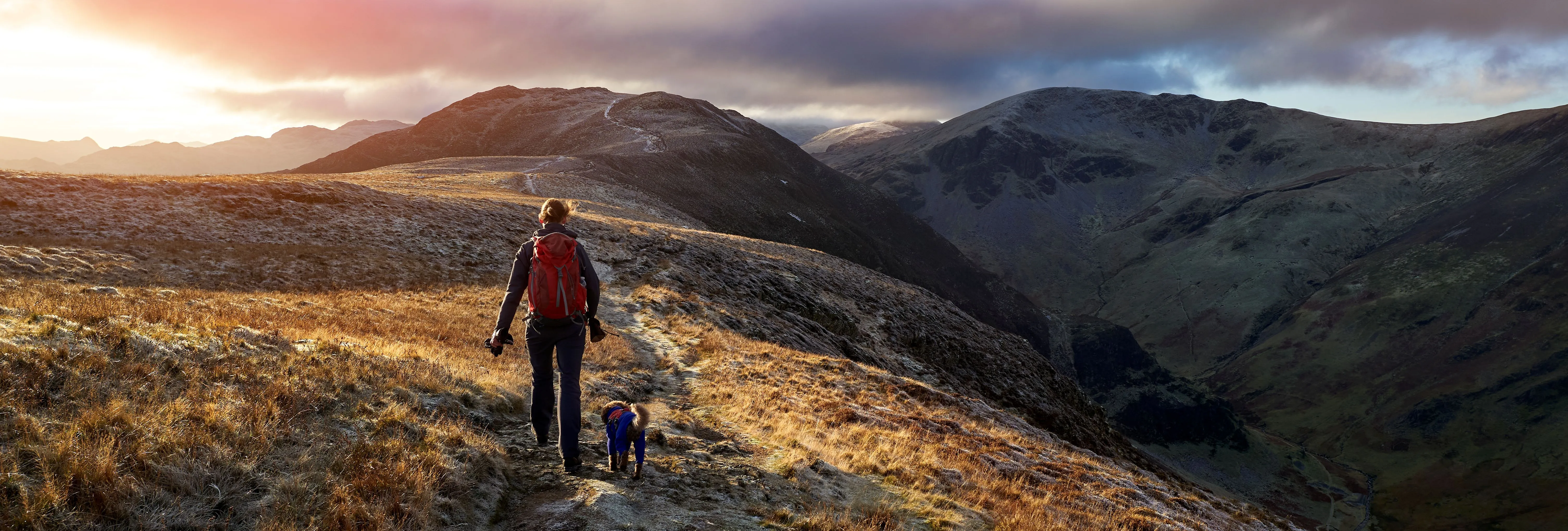 Hiker with a red backpack walking a dog along a frosty mountain ridge at sunrise.