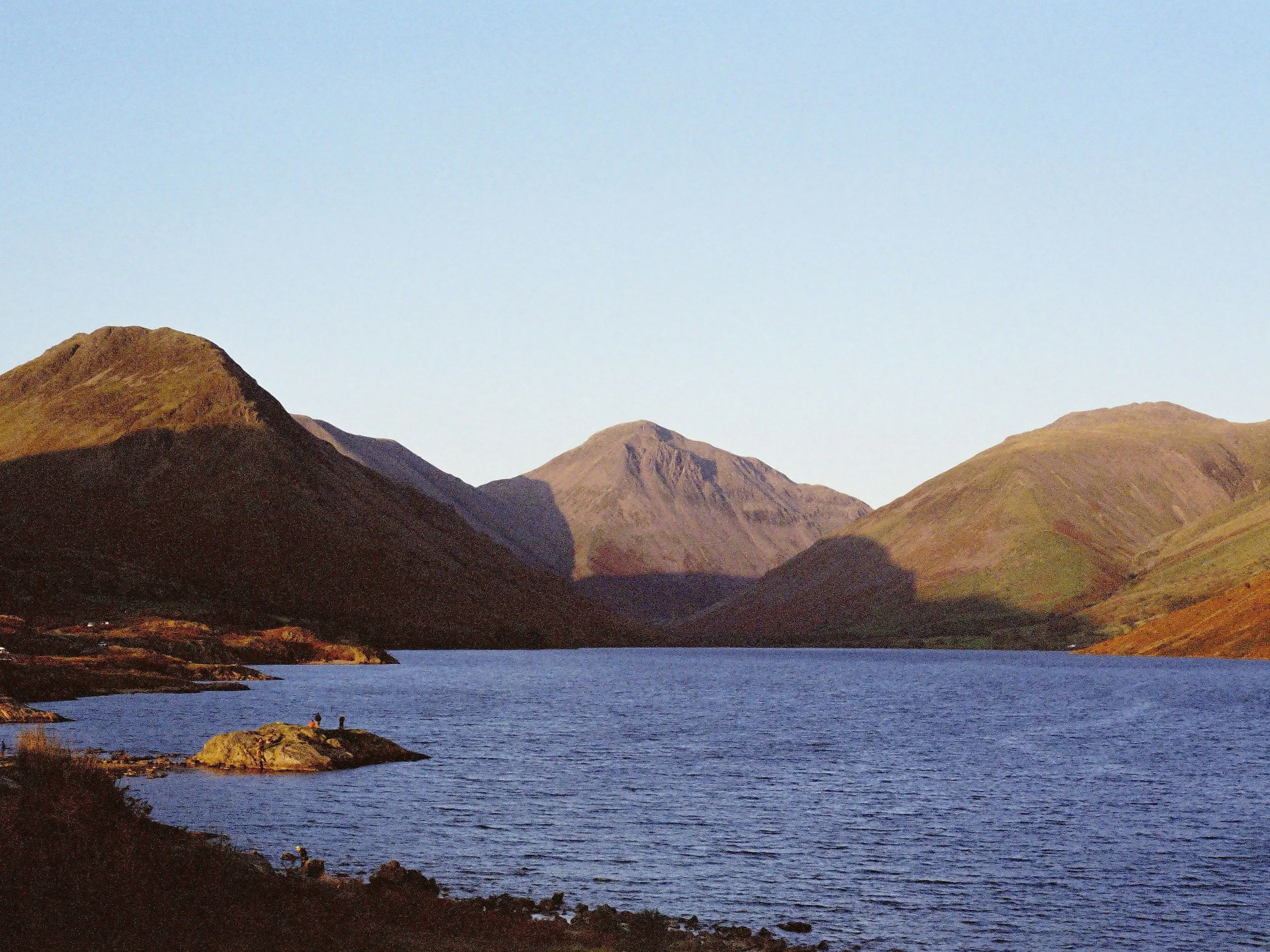 Sunlit mountains surrounding a calm lake beneath a clear pale-blue sky.