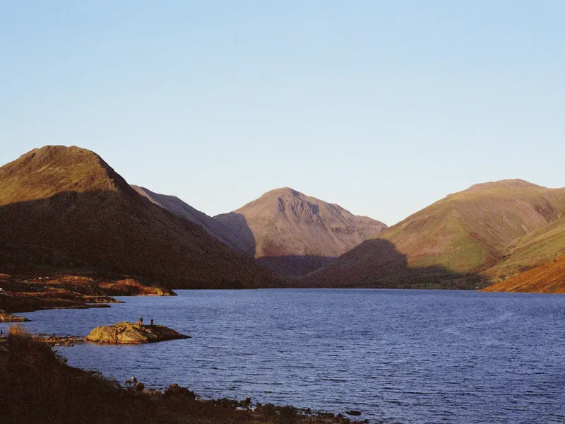 Sunlit mountains surrounding a calm lake beneath a clear pale-blue sky.