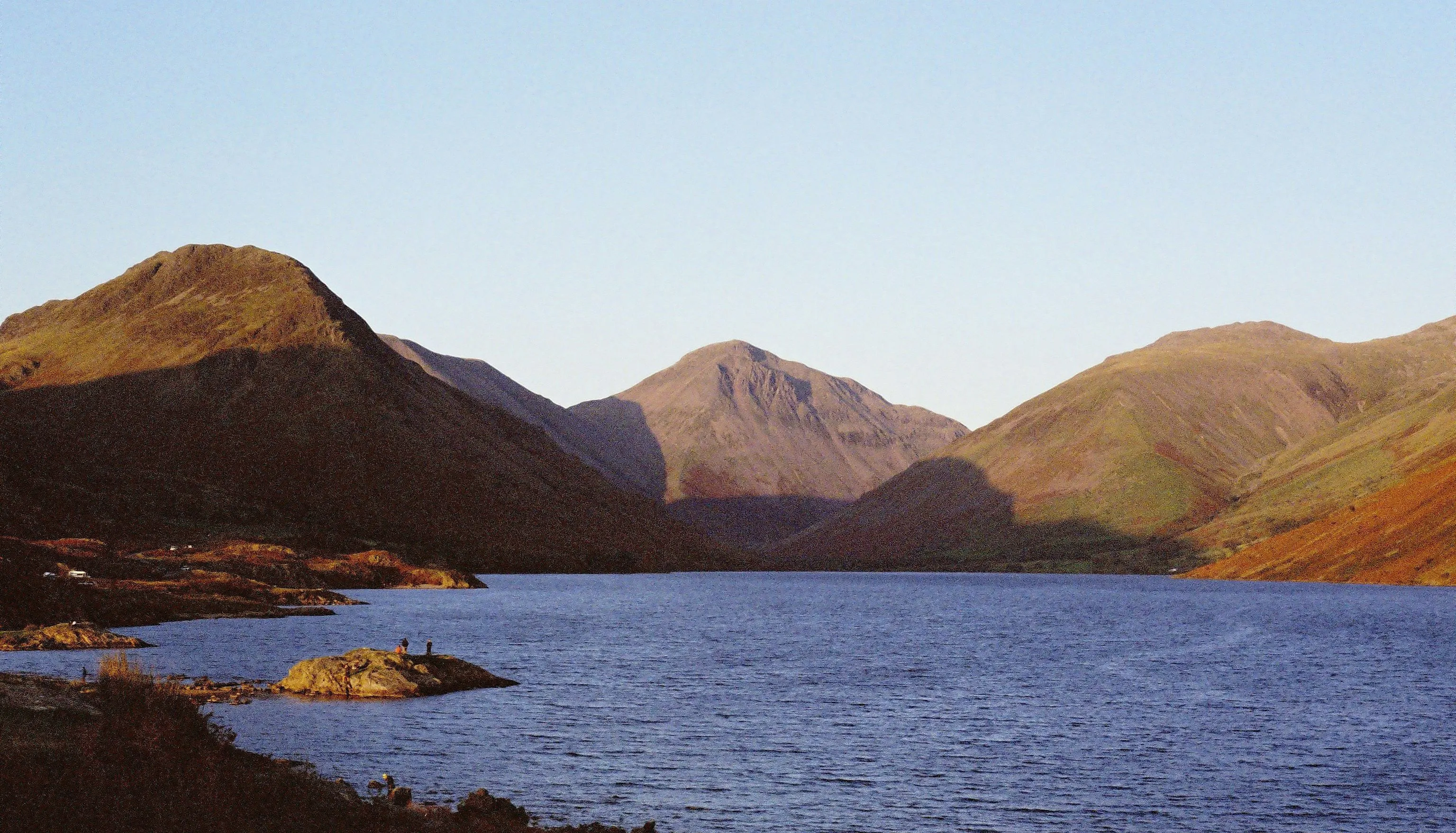 Sunlit mountains surrounding a calm lake beneath a clear pale-blue sky.