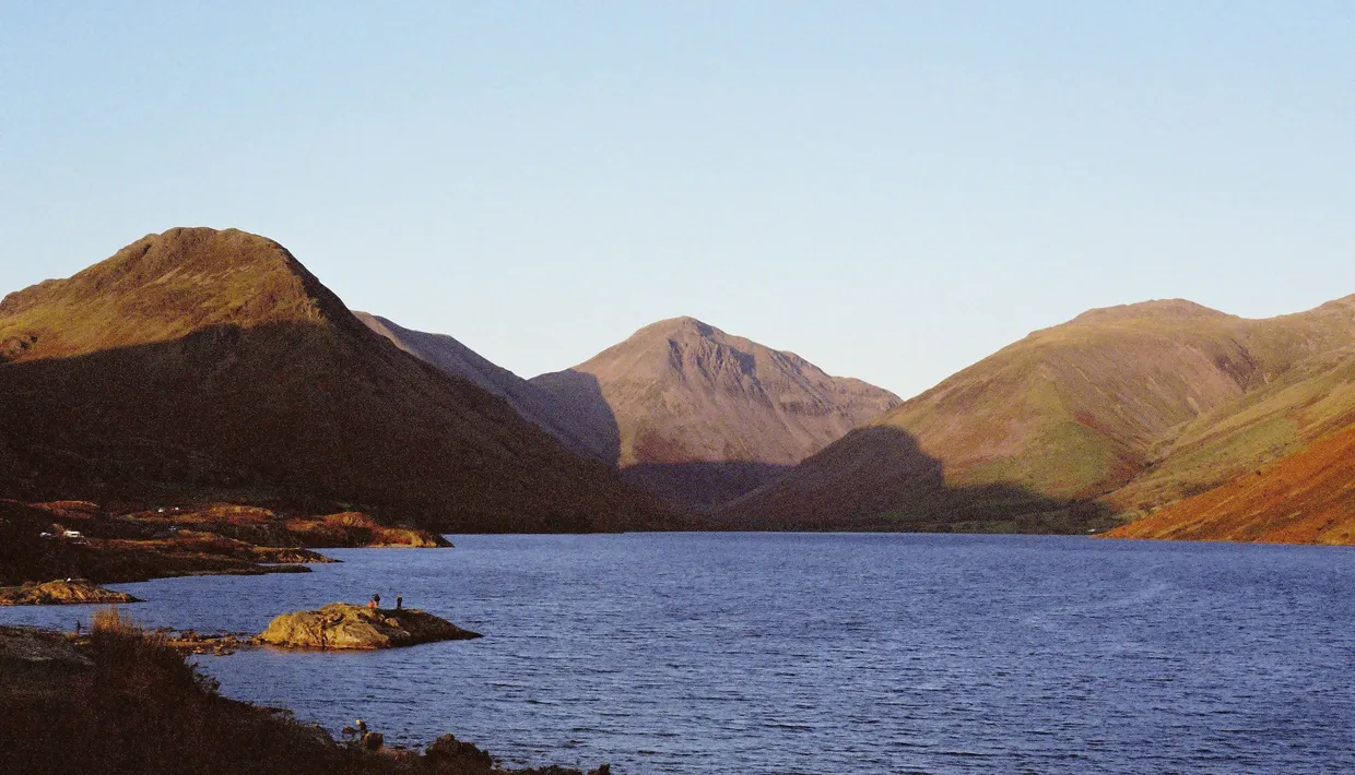 Sunlit mountains surrounding a calm lake beneath a clear pale-blue sky.