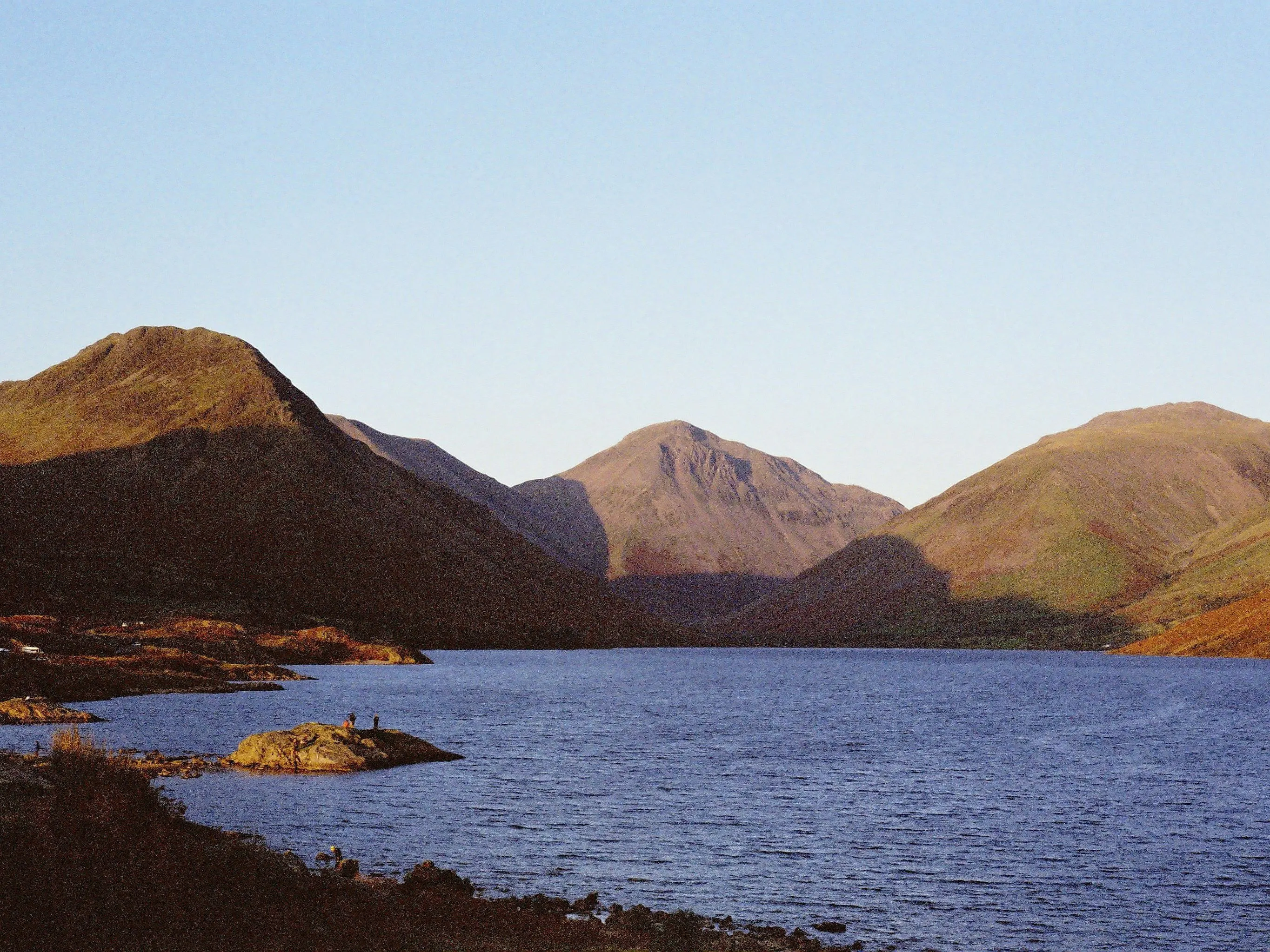 Sunlit mountains surrounding a calm lake beneath a clear pale-blue sky.