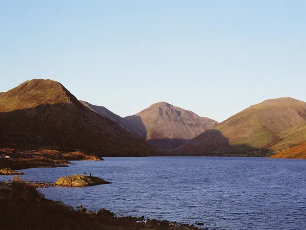 Sunlit mountains surrounding a calm lake beneath a clear pale-blue sky.