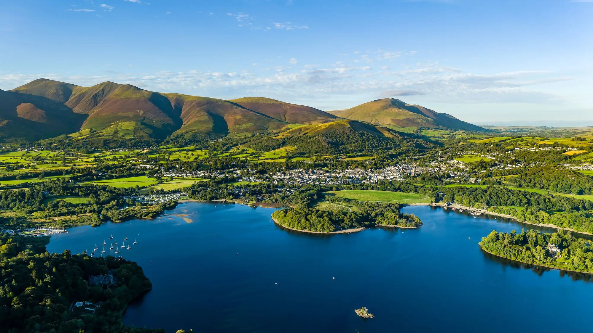 Aerial view of Derwent water lake, Keswick and the fells of the Lake District under a clear blue sky.