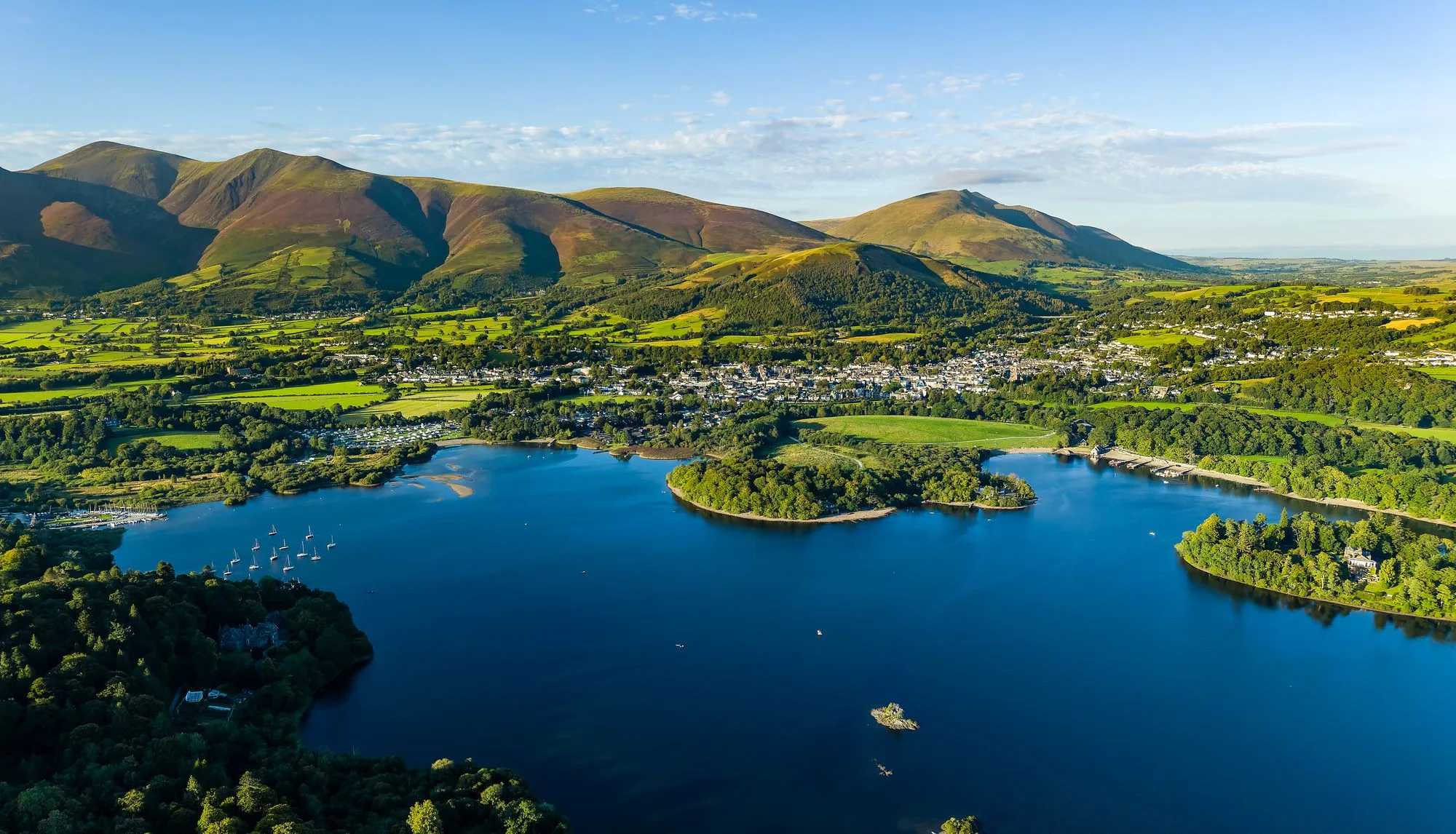 Aerial view of Derwent water lake, Keswick and the fells of the Lake District under a clear blue sky.