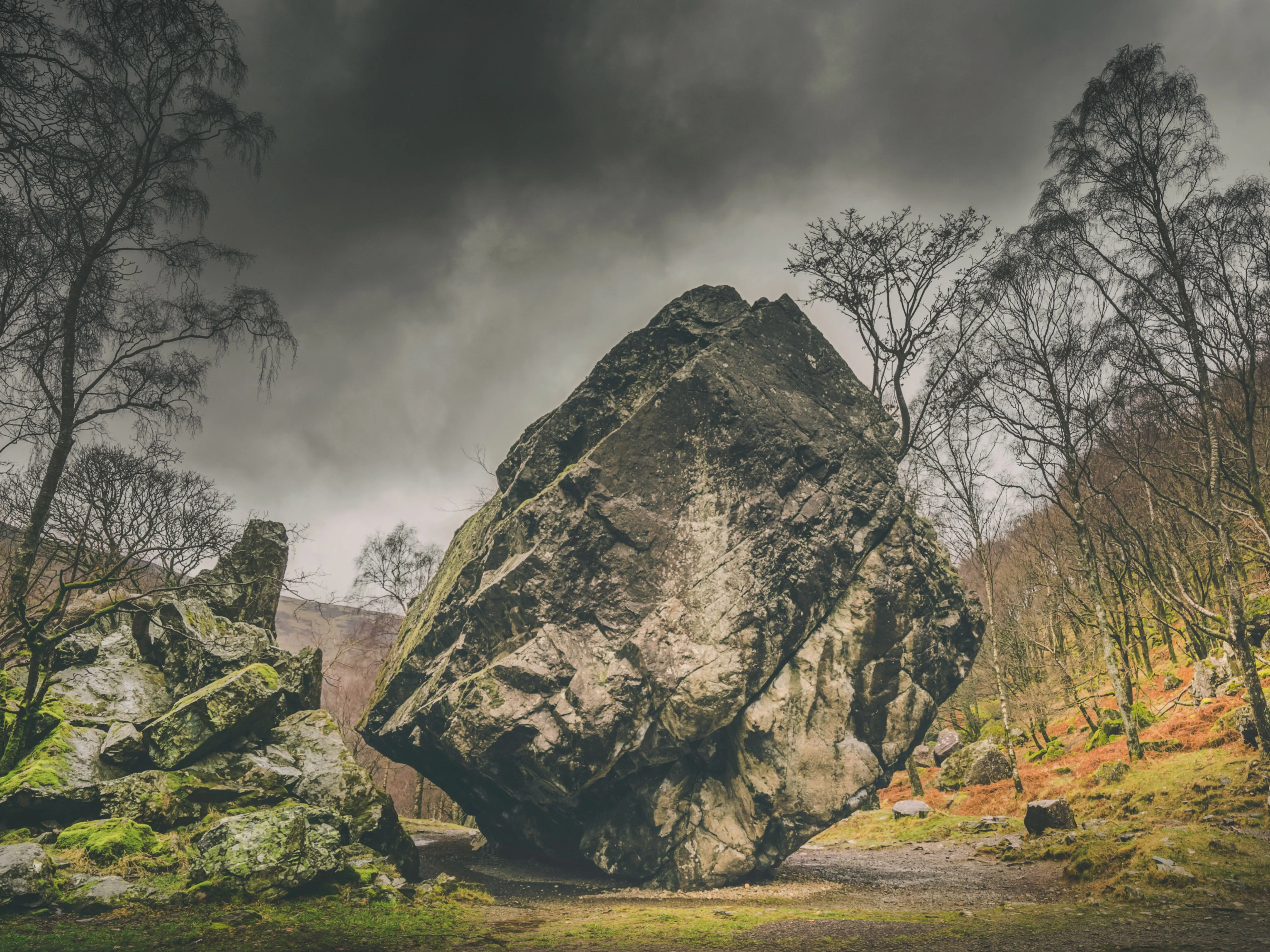 Massive weathered boulder standing among moss-covered rocks and bare trees beneath a dramatic cloudy sky