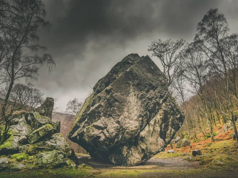 Massive weathered boulder standing among moss-covered rocks and bare trees beneath a dramatic cloudy sky