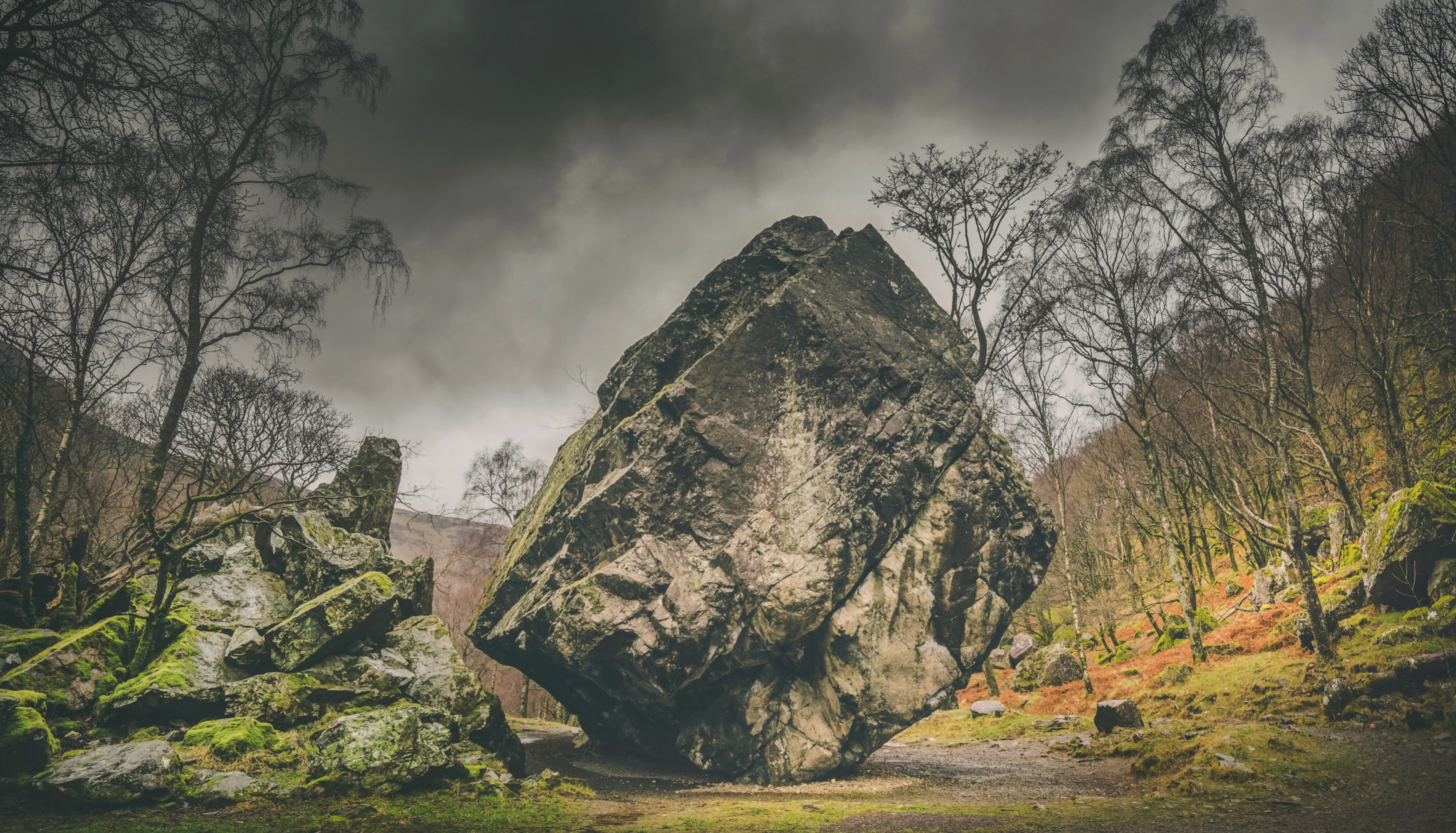 Massive weathered boulder standing among moss-covered rocks and bare trees beneath a dramatic cloudy sky