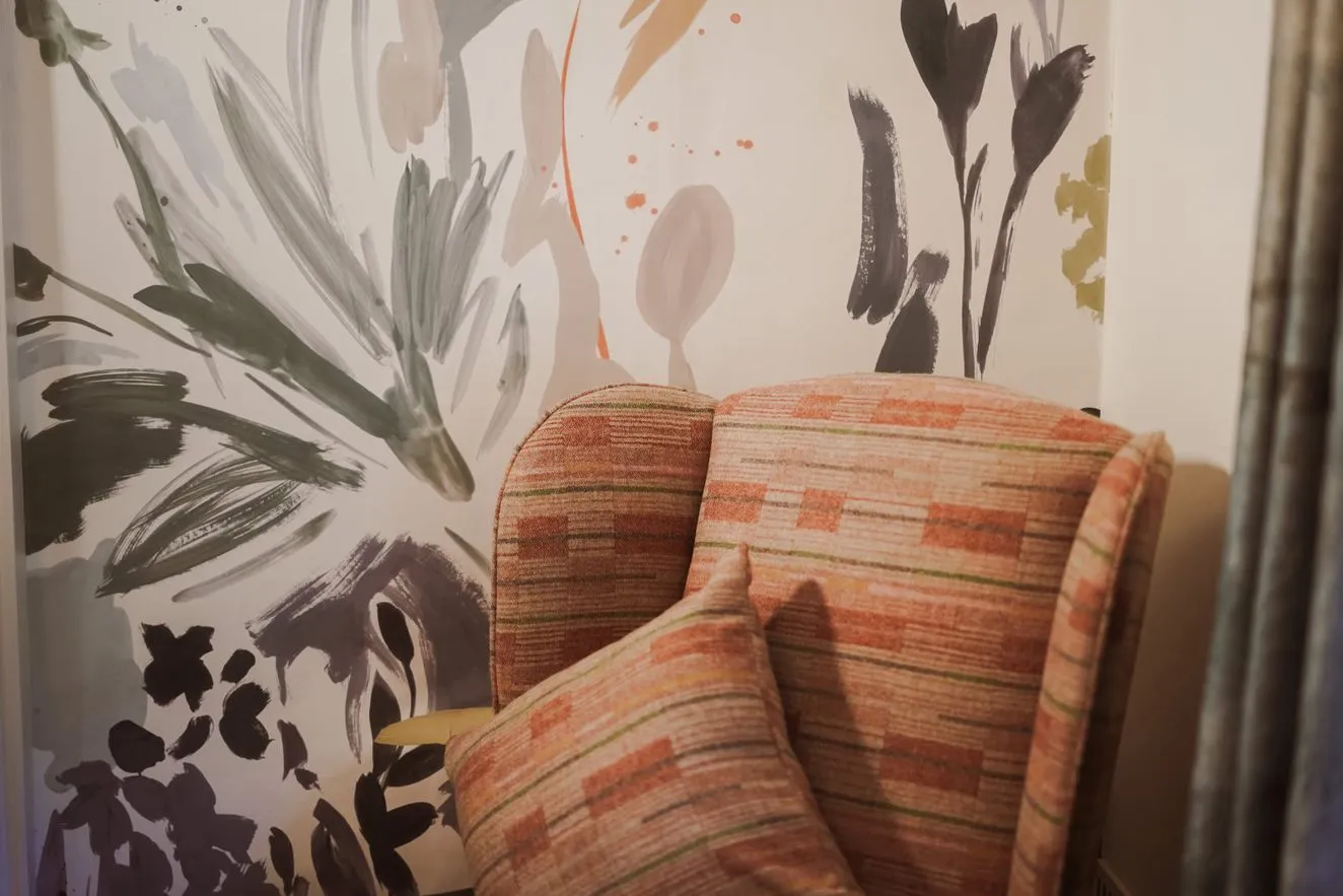 Patterned upholstered armchair with a matching cushion against a wall covered in abstract floral brushstroke wallpaper.