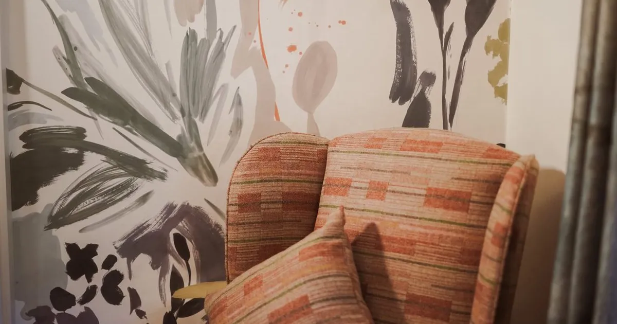 Patterned upholstered armchair with a matching cushion against a wall covered in abstract floral brushstroke wallpaper.