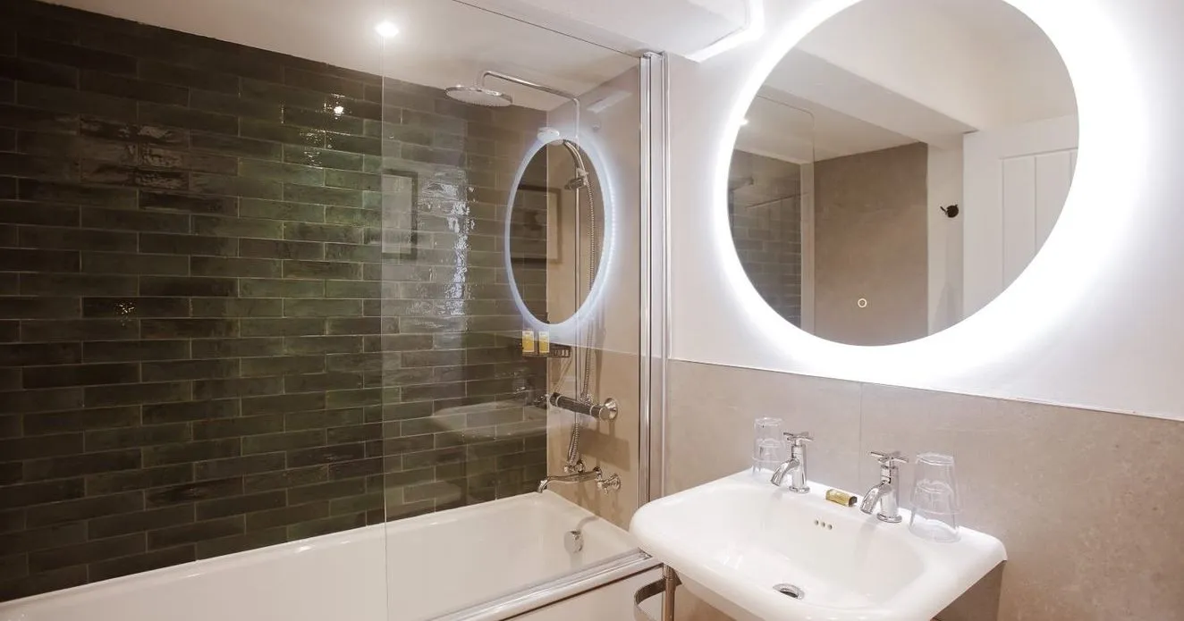 Modern bathroom with a white bath, glass shower screen, dark green subway tiles, pedestal sink and illuminated round mirror.