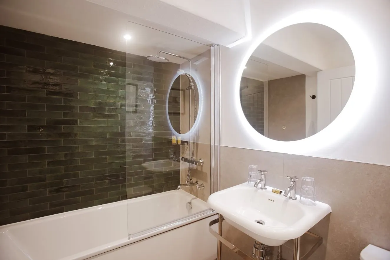 Modern bathroom with a white bath, glass shower screen, dark green subway tiles, pedestal sink and illuminated round mirror.