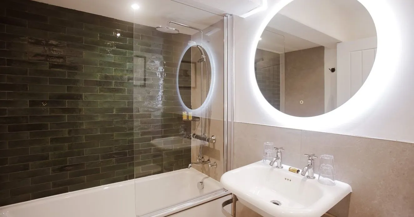 Modern bathroom with a white bath, glass shower screen, dark green subway tiles, pedestal sink and illuminated round mirror.
