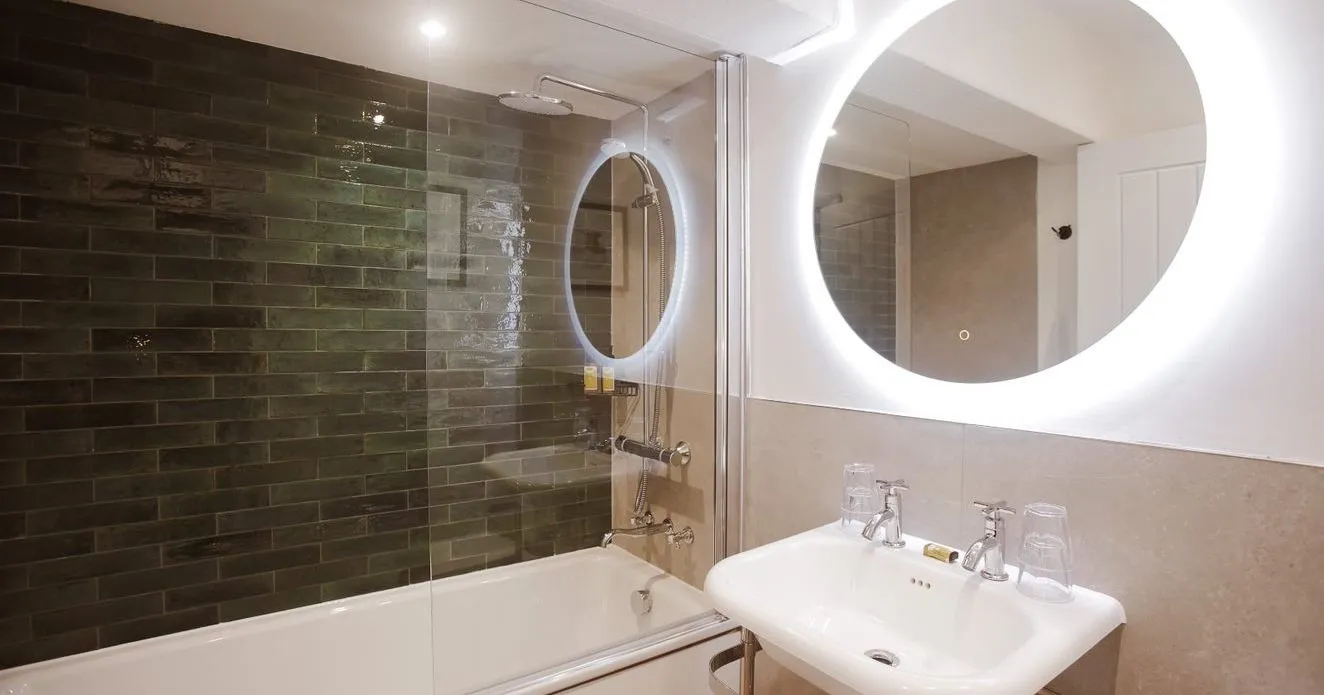 Modern bathroom with a white bath, glass shower screen, dark green subway tiles, pedestal sink and illuminated round mirror.