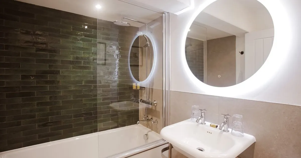 Modern bathroom with a white bath, glass shower screen, dark green subway tiles, pedestal sink and illuminated round mirror.