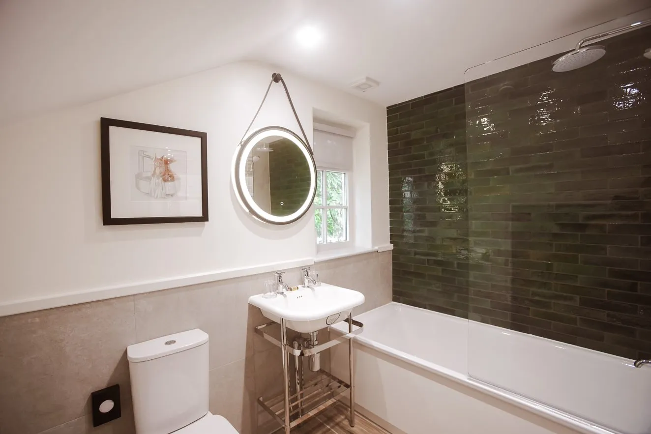 Modern bathroom with a white bath, toilet, pedestal sink and illuminated round mirror beside dark green tiled shower walls.