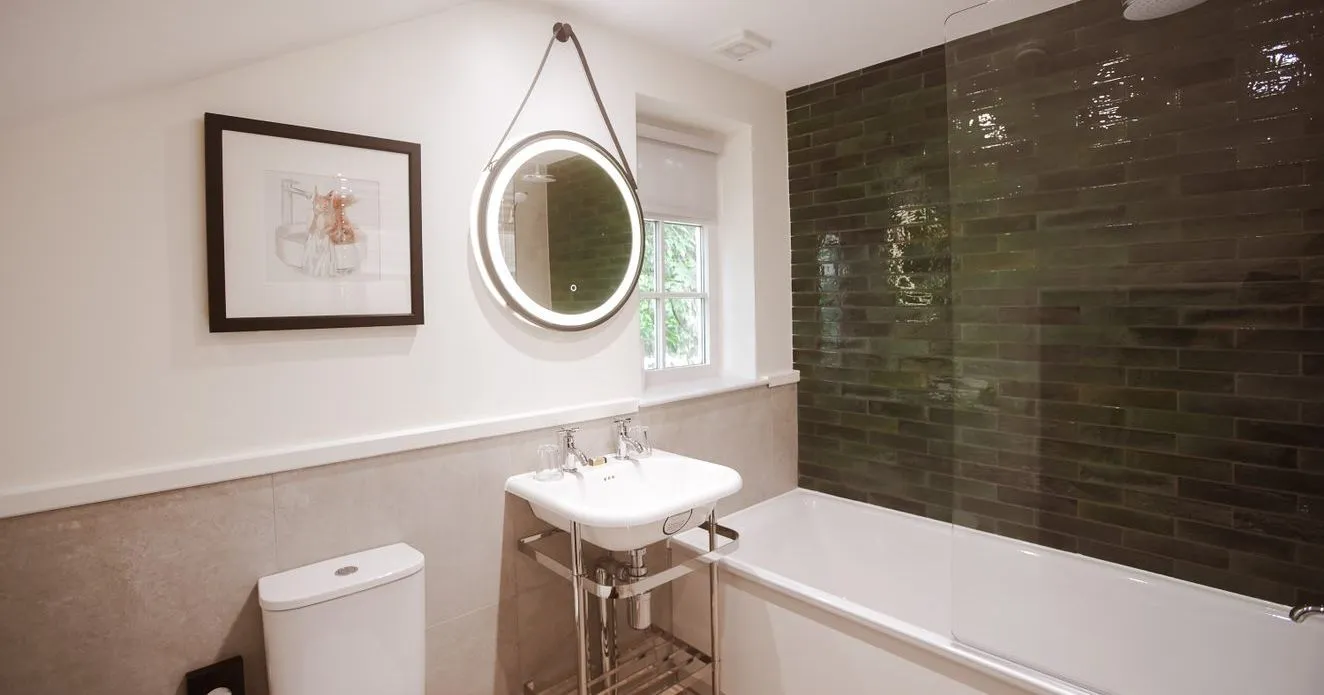 Modern bathroom with a white bath, toilet, pedestal sink and illuminated round mirror beside dark green tiled shower walls.