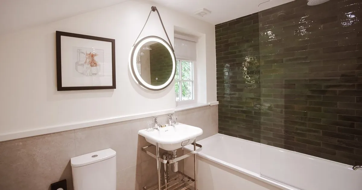 Modern bathroom with a white bath, toilet, pedestal sink and illuminated round mirror beside dark green tiled shower walls.