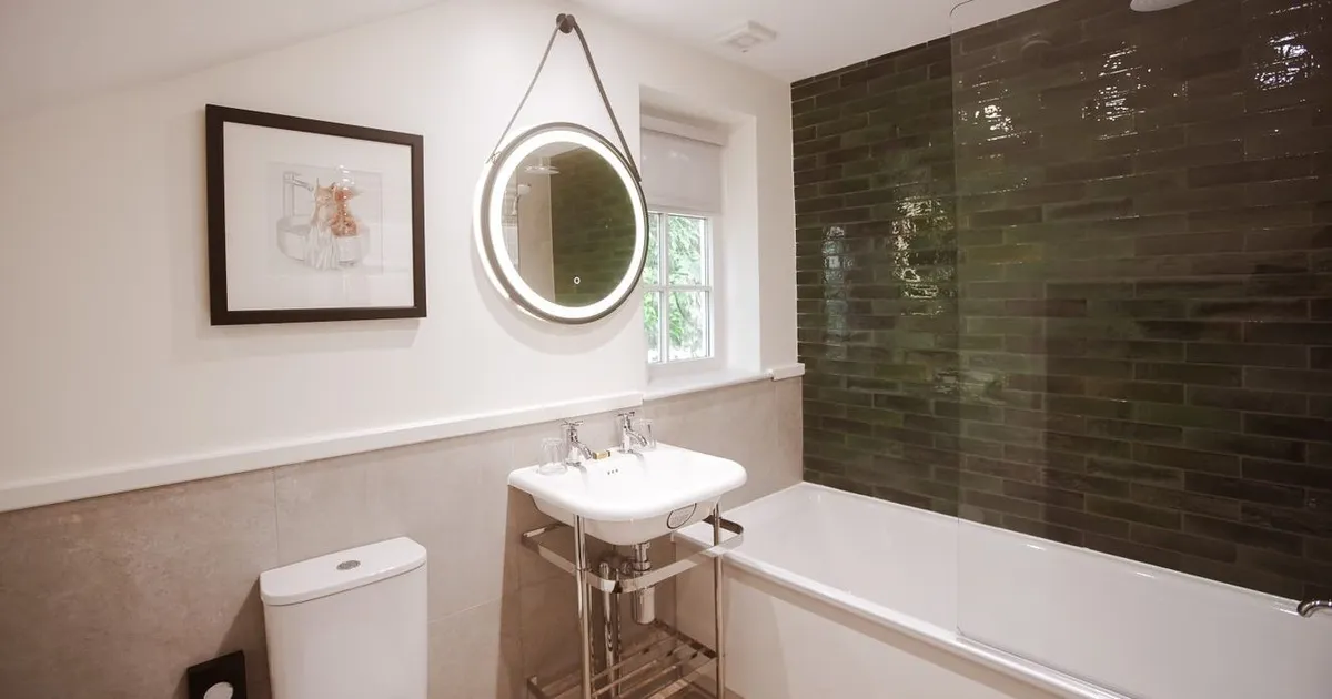 Modern bathroom with a white bath, toilet, pedestal sink and illuminated round mirror beside dark green tiled shower walls.