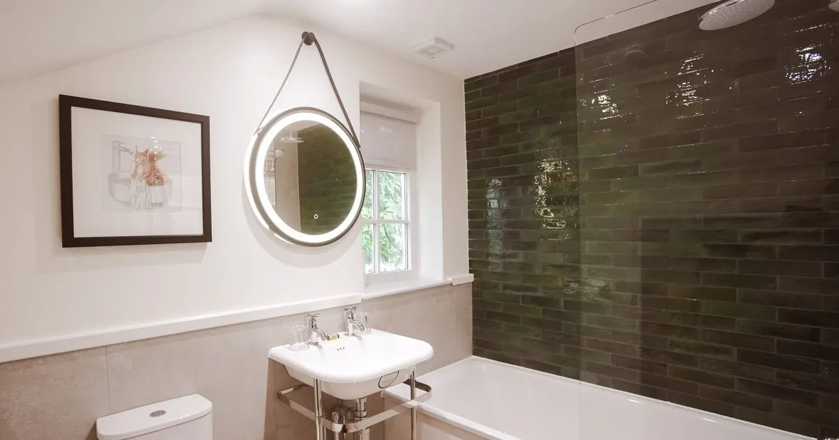 Modern bathroom with a white bath, toilet, pedestal sink and illuminated round mirror beside dark green tiled shower walls.