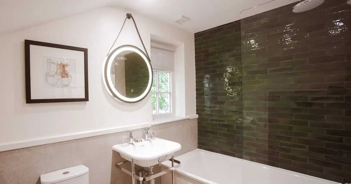 Modern bathroom with a white bath, toilet, pedestal sink and illuminated round mirror beside dark green tiled shower walls.