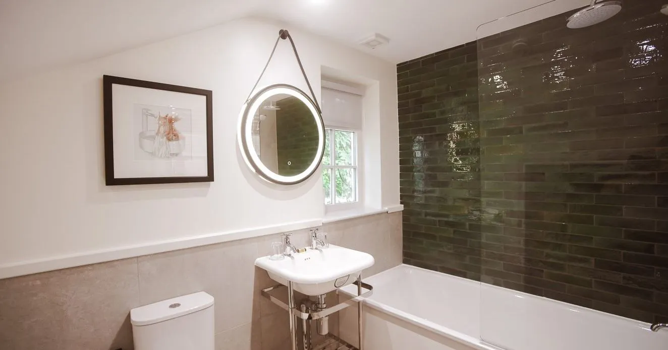 Modern bathroom with a white bath, toilet, pedestal sink and illuminated round mirror beside dark green tiled shower walls.