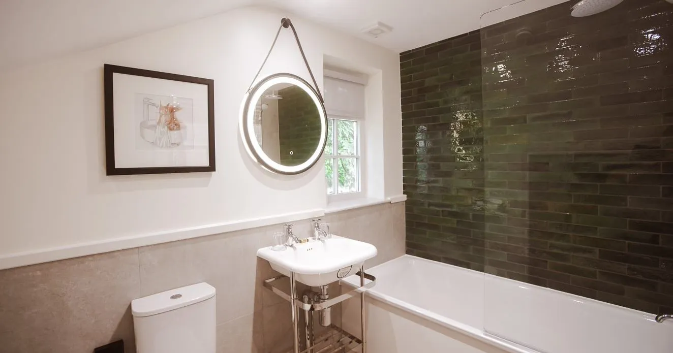 Modern bathroom with a white bath, toilet, pedestal sink and illuminated round mirror beside dark green tiled shower walls.