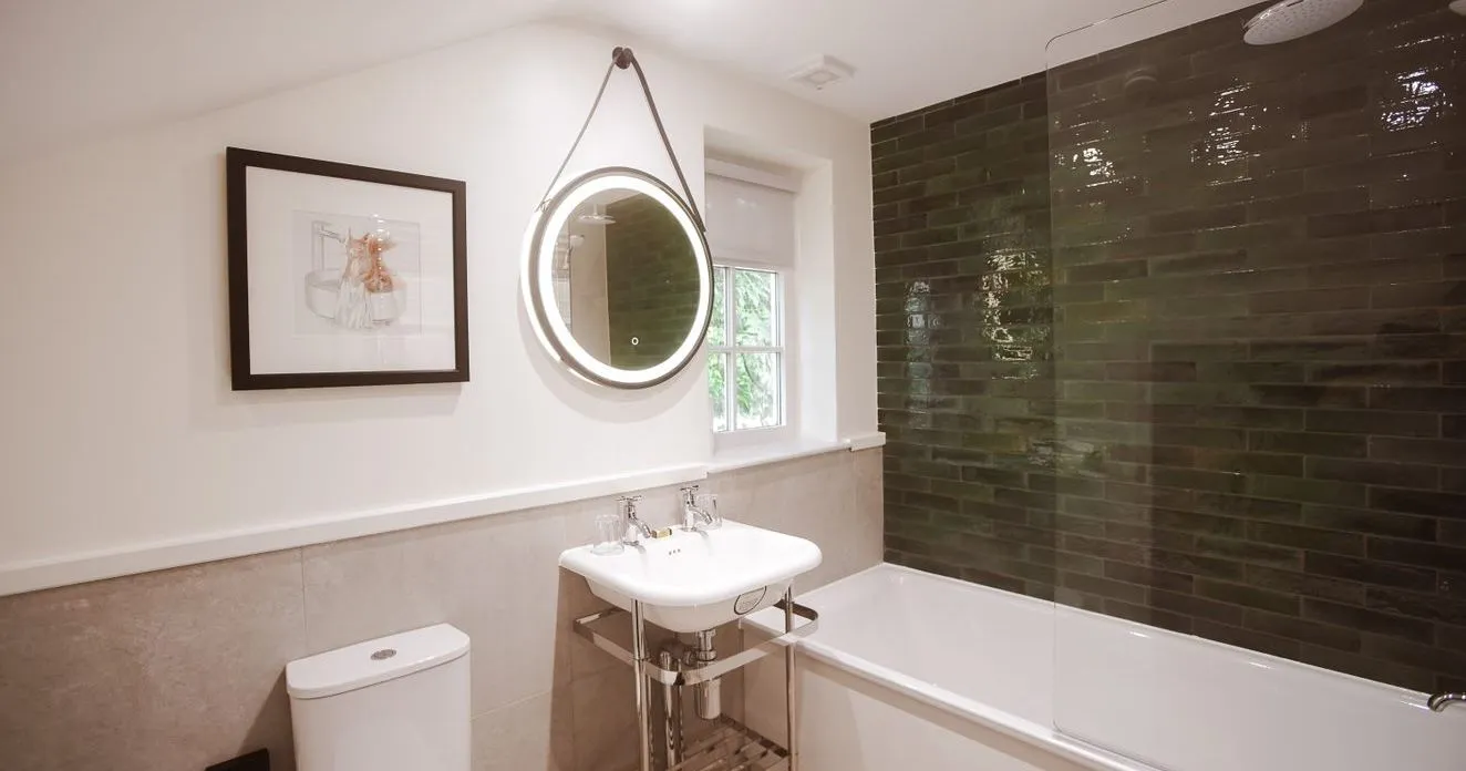 Modern bathroom with a white bath, toilet, pedestal sink and illuminated round mirror beside dark green tiled shower walls.