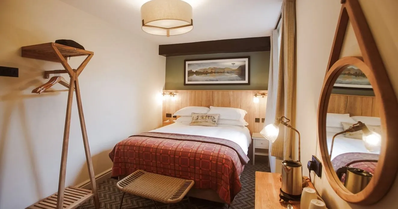 Cosy hotel bedroom with a double bed, red patterned throw, wooden luggage rack and round mirror.