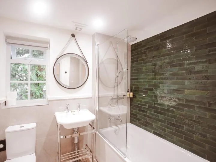 Bright modern bathroom with a white bathtub, glass shower screen, green tiled feature wall and round hanging mirror.