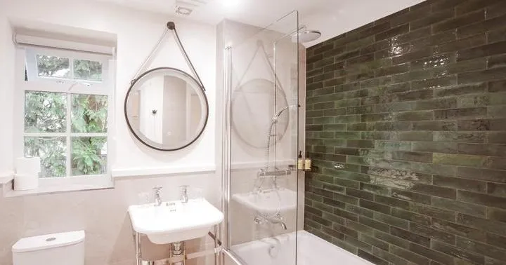 Bright modern bathroom with a white bathtub, glass shower screen, green tiled feature wall and round hanging mirror.