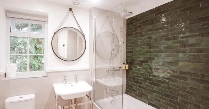 Bright modern bathroom with a white bathtub, glass shower screen, green tiled feature wall and round hanging mirror.